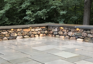 Landscape design by Nest Outdoors featuring a custom stone patio with a sitting wall and landscape lighting
